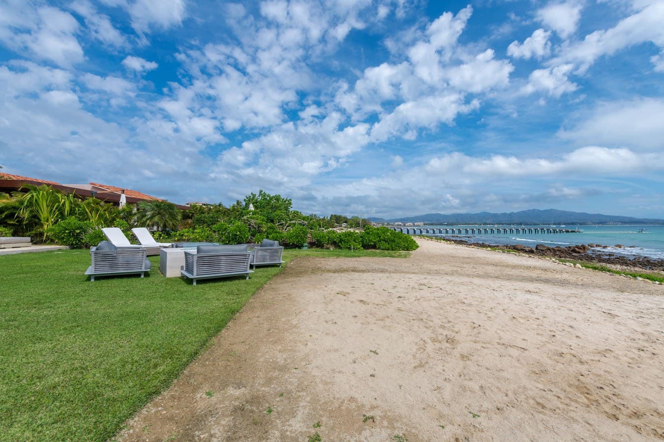 This beachfront property offers direct sandy beach access with lush lawn space and loungers overlooking the ocean. The distinctive arched bridge stretches across the calm bay waters, framing the mountain backdrop.