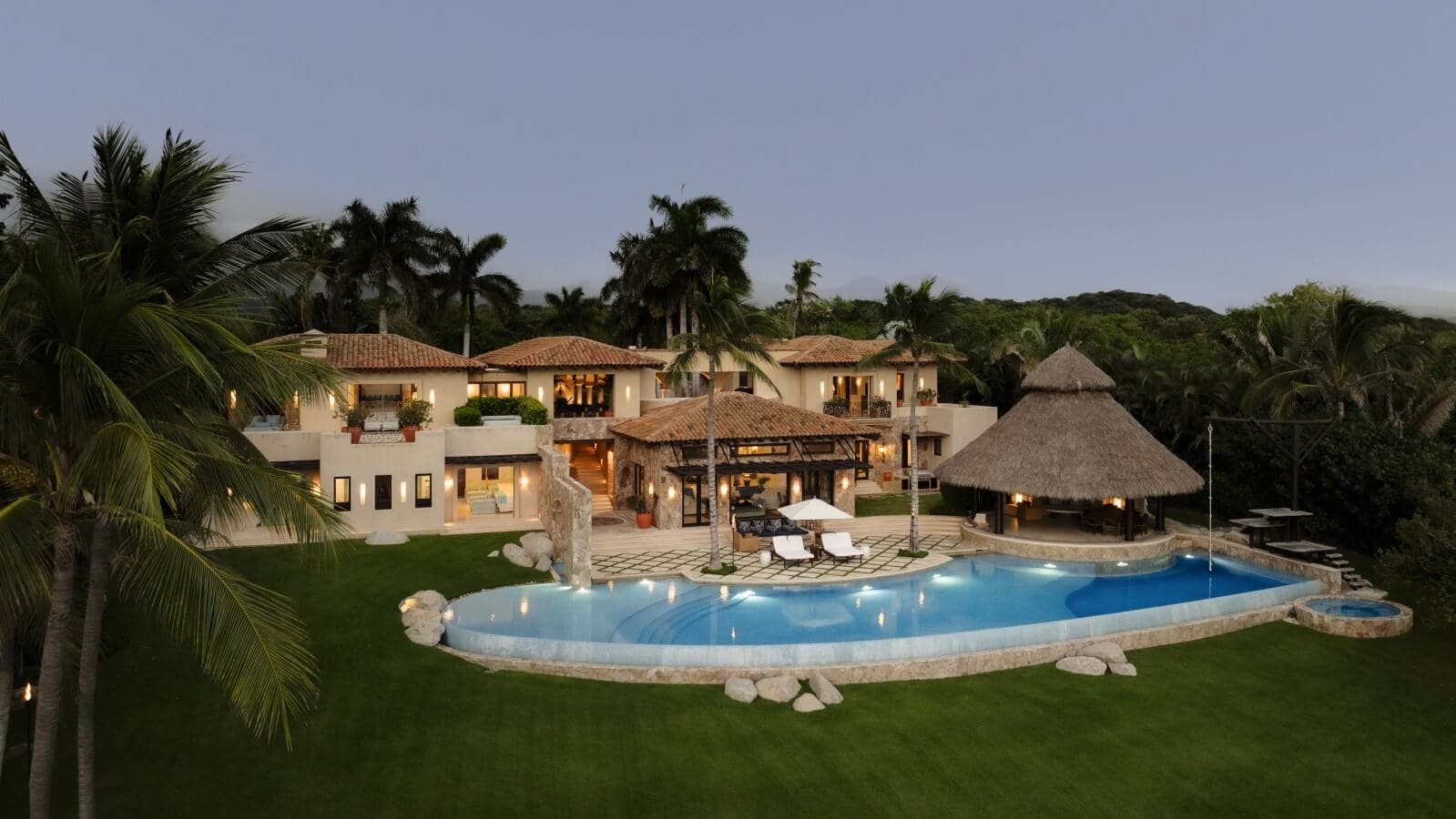 A stunning tropical estate nestled among lush palms and verdant hillsides, featuring a sparkling pool and thatched-roof palapa at twilight.