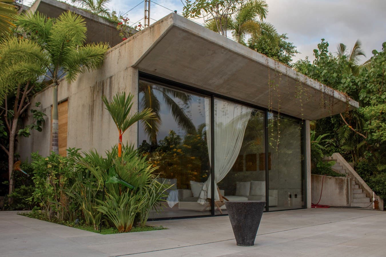 Modern concrete architecture nestled in tropical gardens, where floor-to-ceiling glass walls invite natural light into your private sanctuary surrounded by lush palms and greenery.