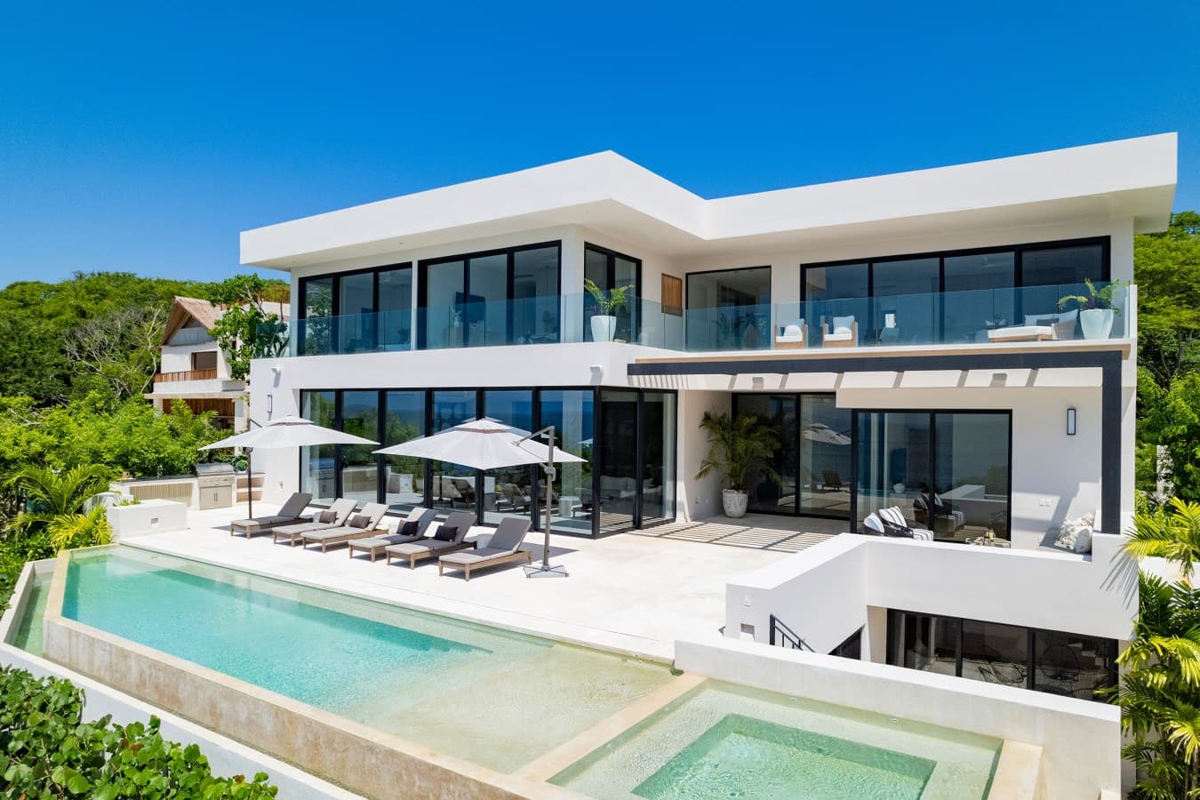 A modern three-level villa nestled in lush tropical surroundings, featuring clean white architecture, expansive glass walls, and a stunning hillside setting with tiered pool decks overlooking the landscape.