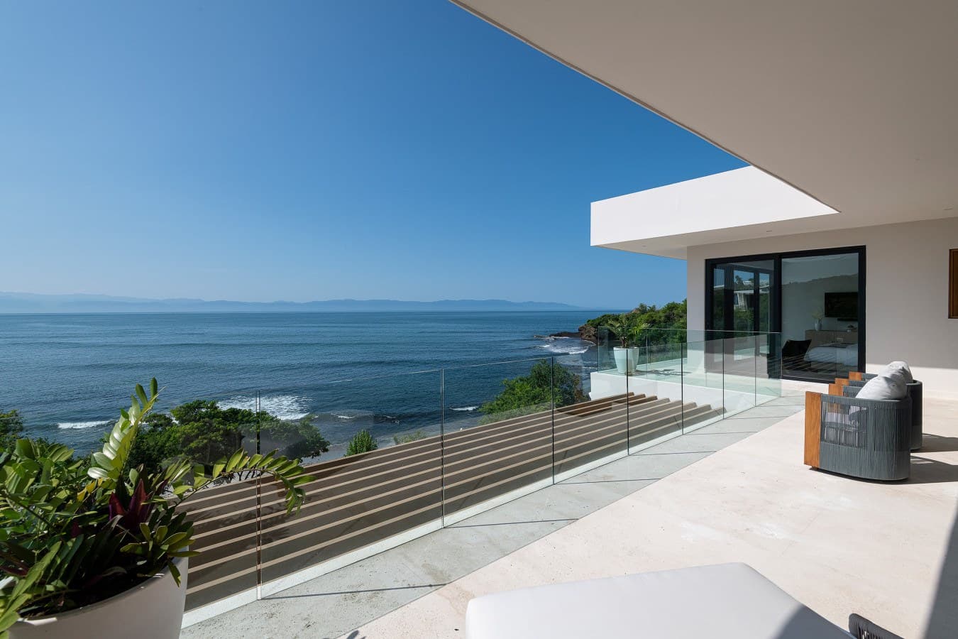 Breathtaking ocean views stretch endlessly from your private terrace, where glass railings frame the shimmering water and coastal greenery below. Sink into modern outdoor seating and let the sea breeze wash over you in this stunning, serene escape.