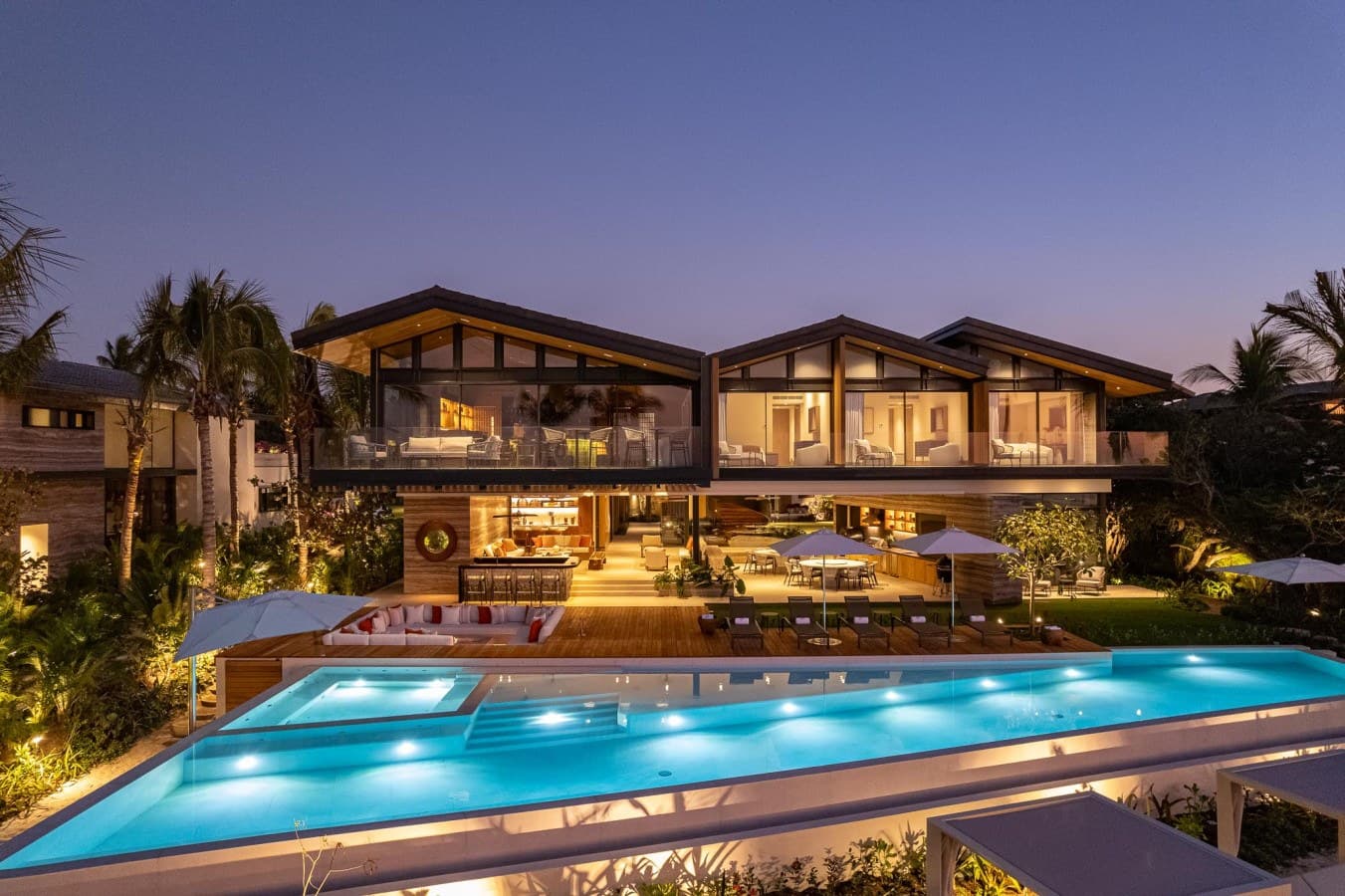 A stunning beachfront villa illuminated at twilight, surrounded by swaying palms and tropical landscaping, with a dramatic blue swimming pool at its heart.