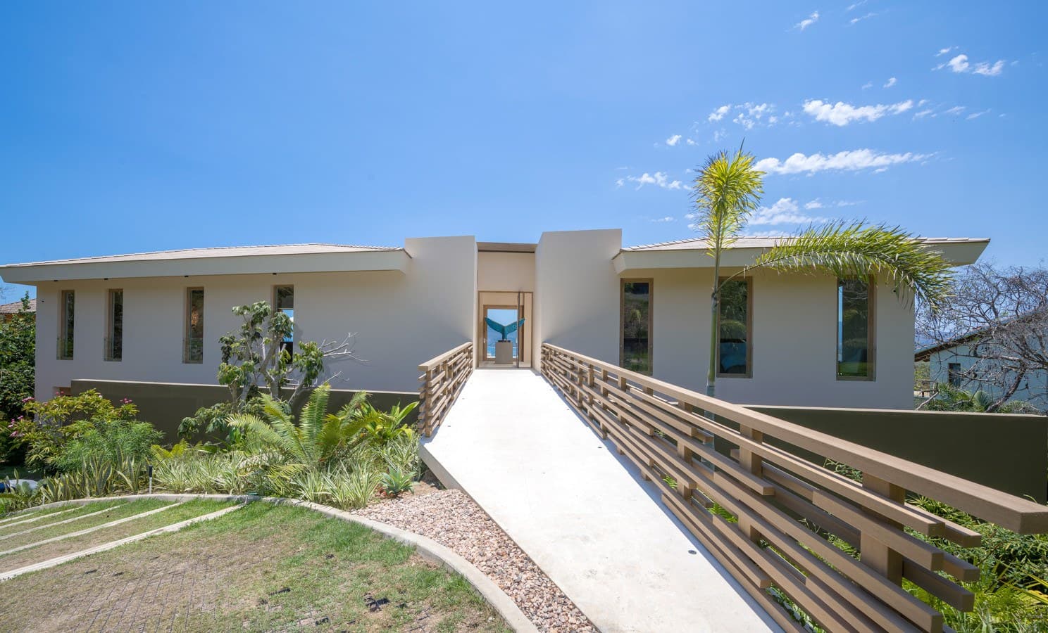 A modern tropical villa awaits, featuring clean architectural lines, lush landscaping, and an inviting walkway leading to the bright entrance under clear blue skies.