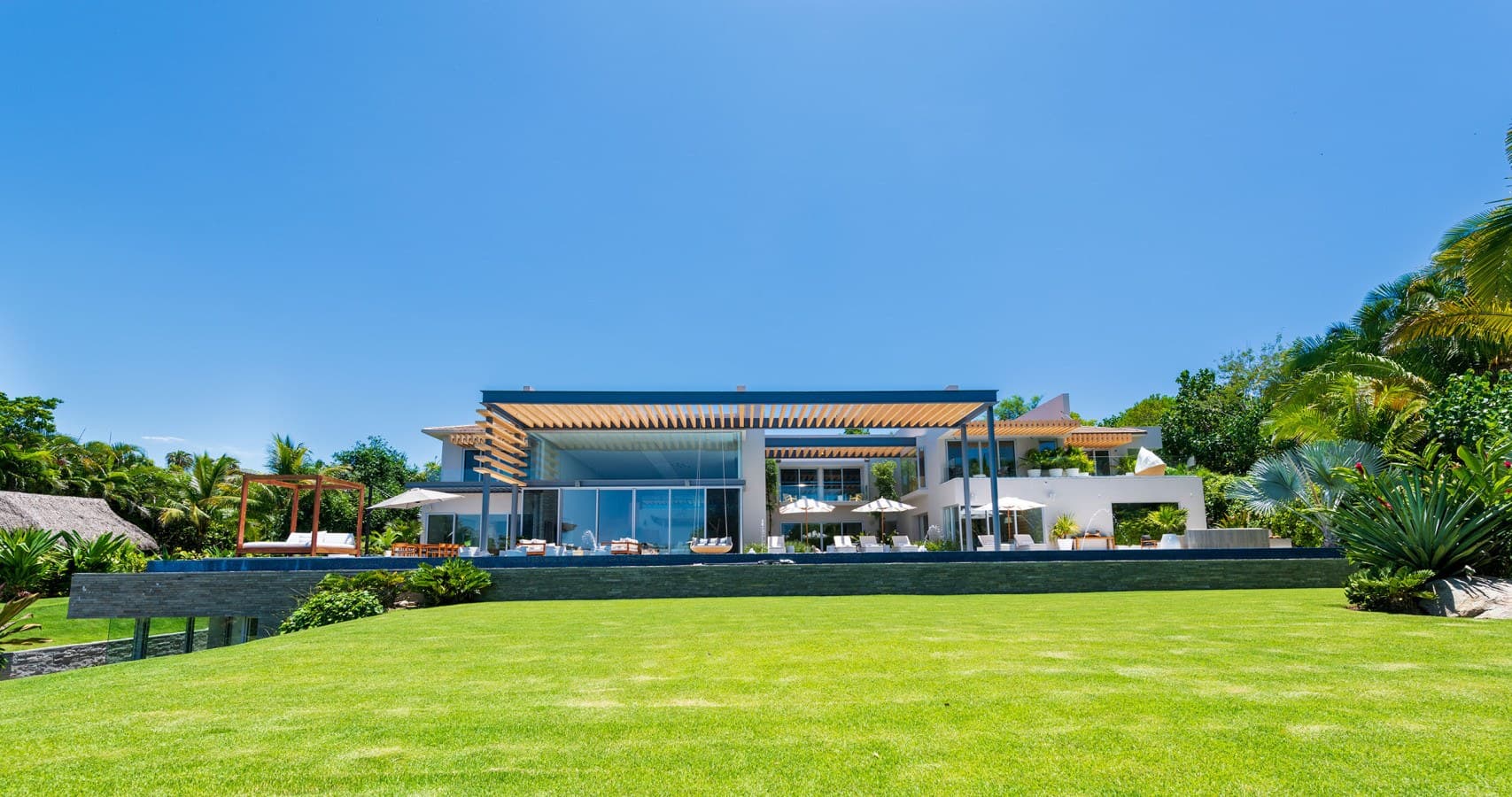 A stunning contemporary estate set within a lush tropical landscape. The expansive manicured lawn and surrounding palm trees frame this architectural showpiece, promising a luxurious private retreat.