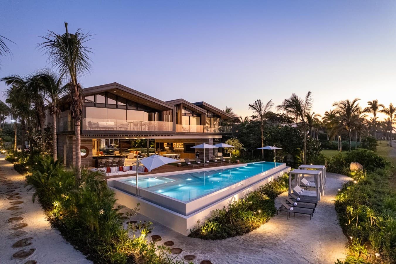 A stunning tropical retreat at dusk, where palm trees sway beside an illuminated infinity-edge pool and contemporary open-air design invites complete relaxation in paradise.