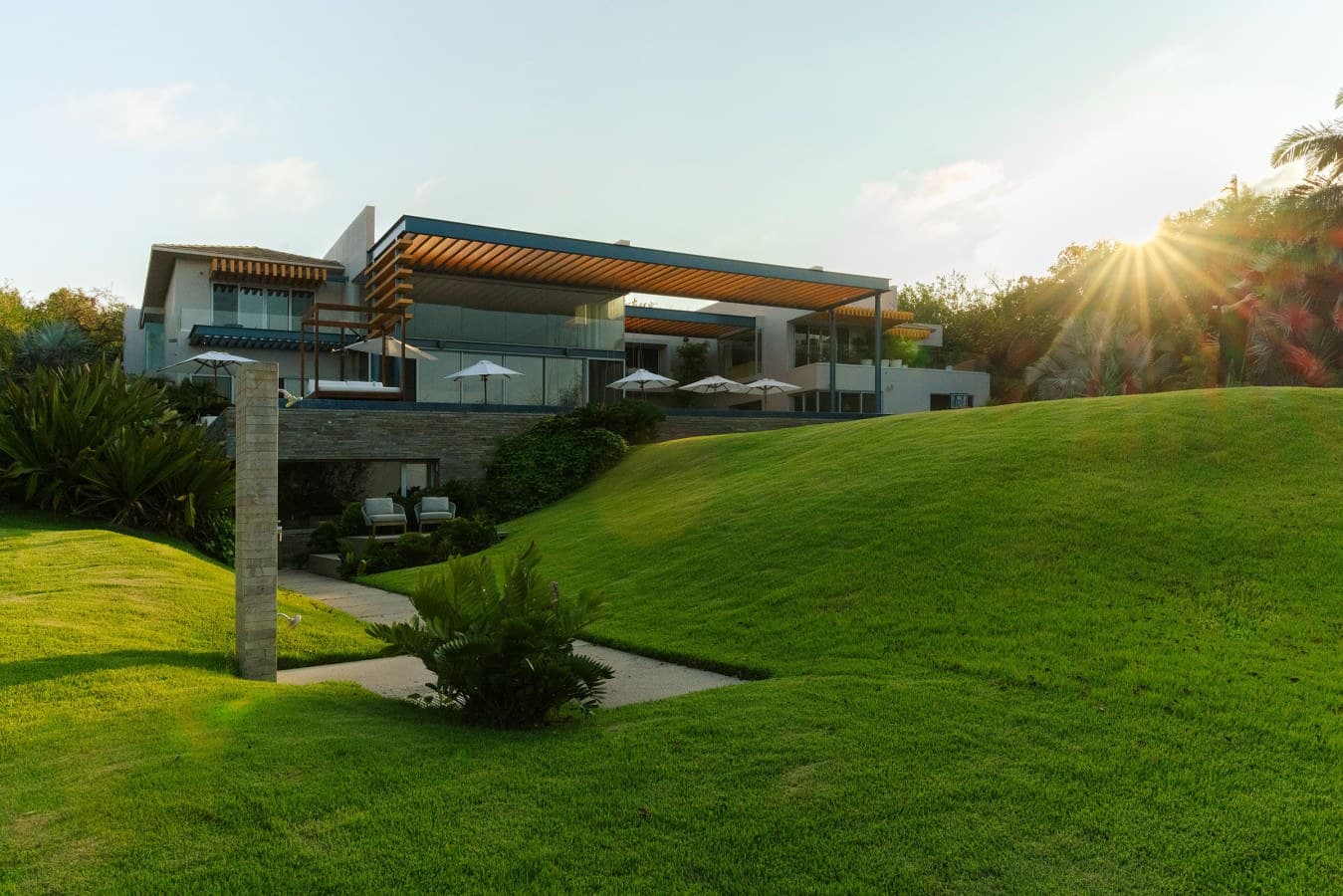 A striking modern property set on a lush, manicured hillside with expansive lawns and mature landscaping, captured at golden hour as the sun sets over the surrounding trees.
