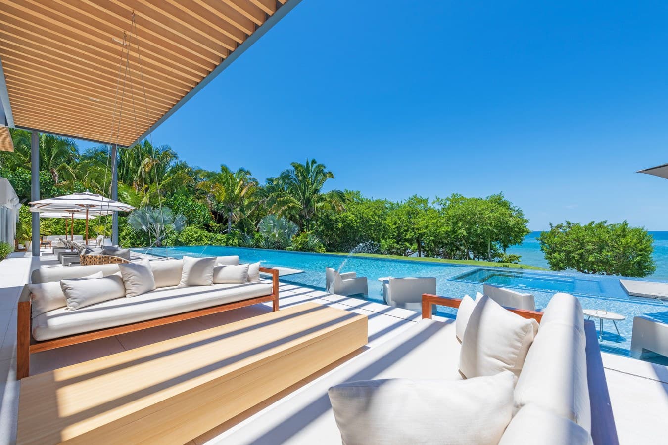 A stunning infinity pool flows seamlessly into turquoise ocean views, framed by lush tropical palms and pristine white lounges under a sleek wooden pergola—your perfect tropical escape.