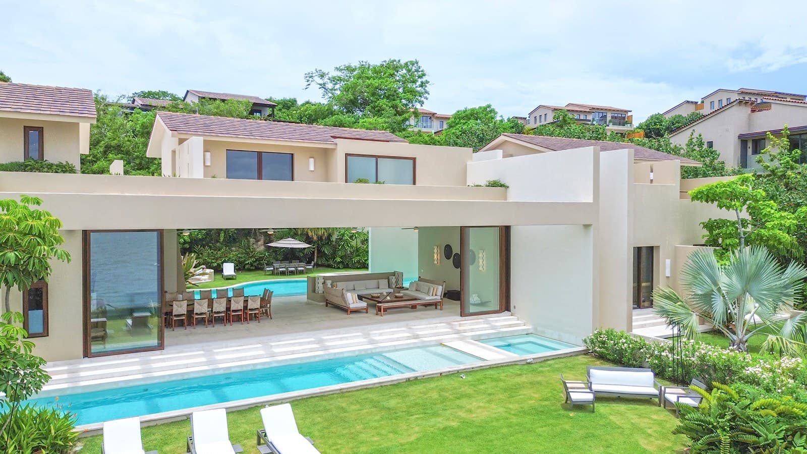 A stunning modern villa nestled among lush tropical landscaping, featuring expansive terraces and multiple pools set against vibrant greenery and neighboring hillside homes.