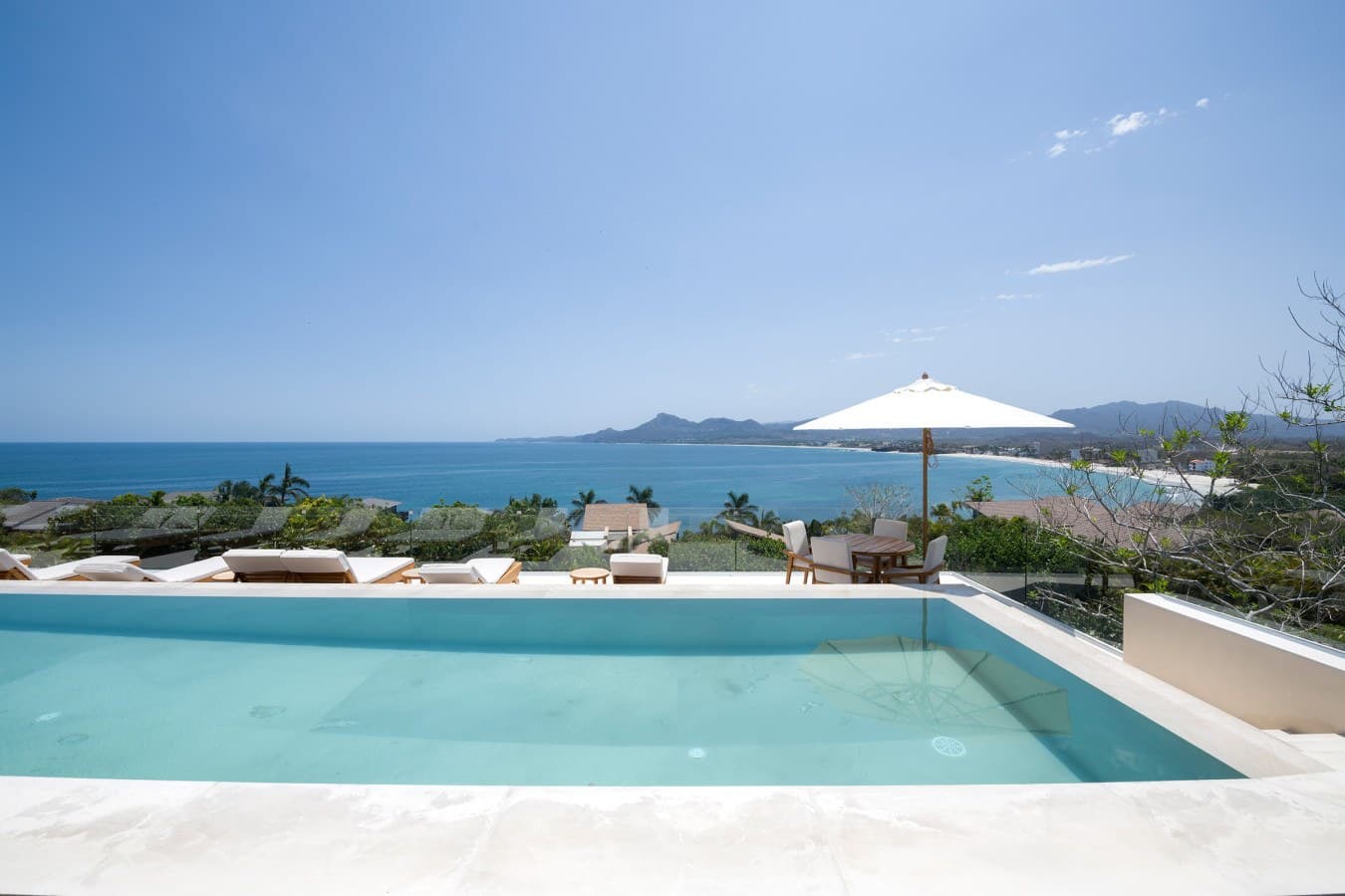 Private infinity pool with panoramic ocean views and distant mountain backdrop, surrounded by lush tropical landscaping and elegant white loungers.