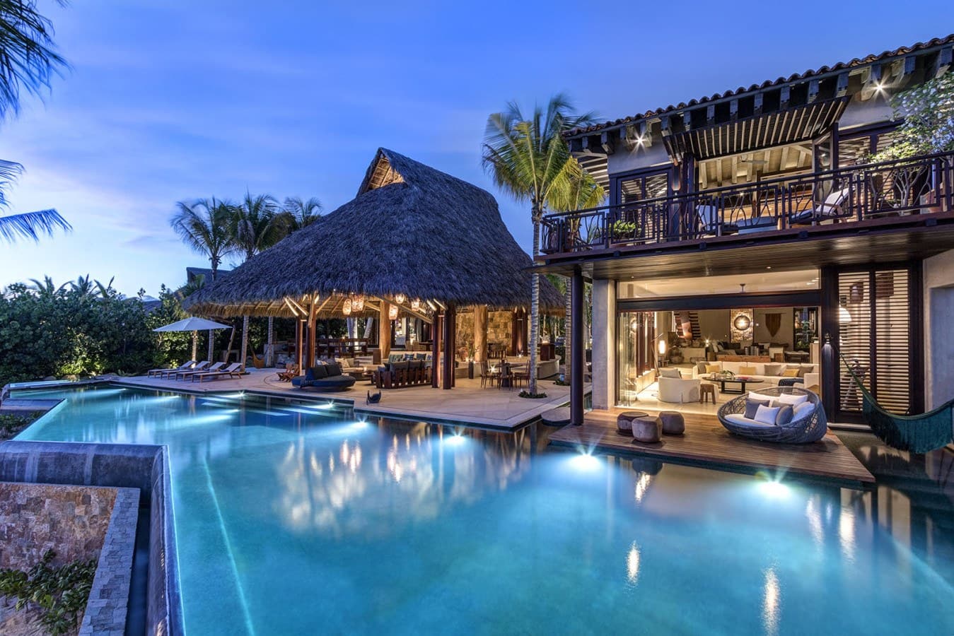 A stunning tropical estate captured at dusk, featuring a thatched-roof palapa, multi-level architecture, and an illuminated infinity pool surrounded by lush palm trees and natural landscaping.