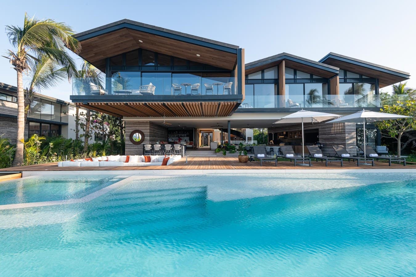 A stunning contemporary tropical villa featuring sleek architecture with floor-to-ceiling windows, warm wood accents, and an inviting infinity pool surrounded by palm trees and modern loungers.