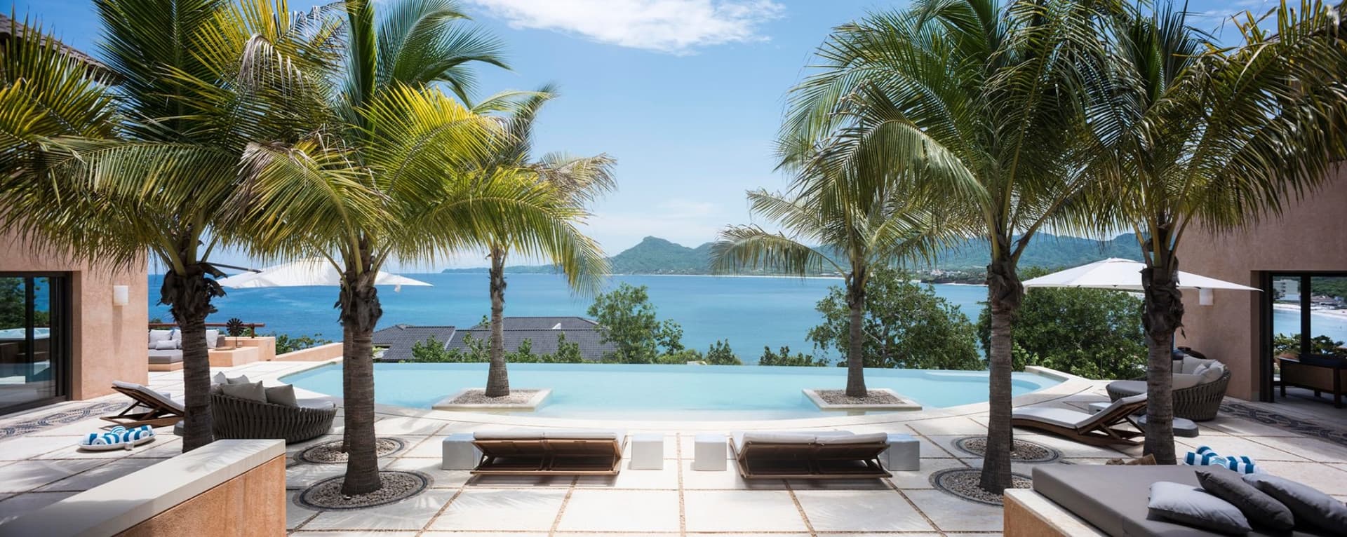 Stunning oceanfront infinity pool surrounded by swaying palms, with pristine blue waters and mountain vistas stretching beyond.