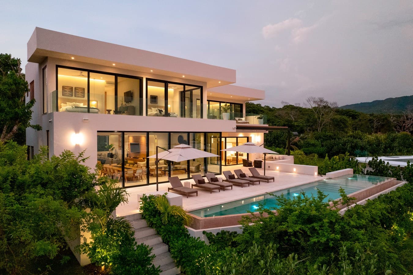 Modern luxury villa with private pool and mountain views, nestled in lush tropical surroundings and illuminated for a serene evening atmosphere.