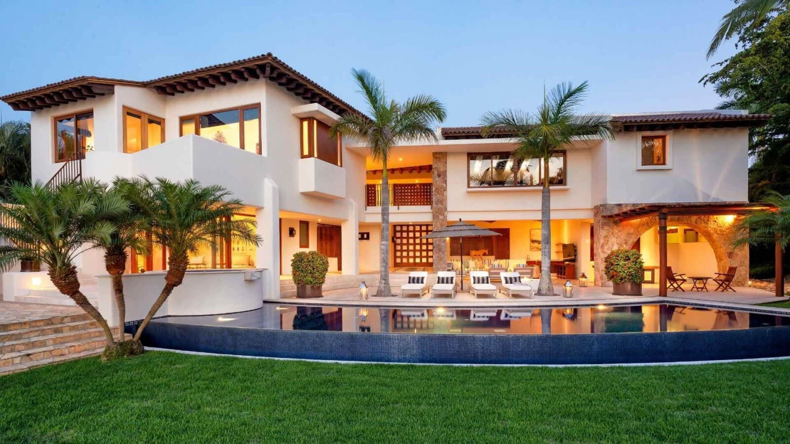 A stunning Spanish-style villa surrounded by lush tropical gardens and an elegant infinity pool, captured during golden hour as warm lights begin to glow from within.