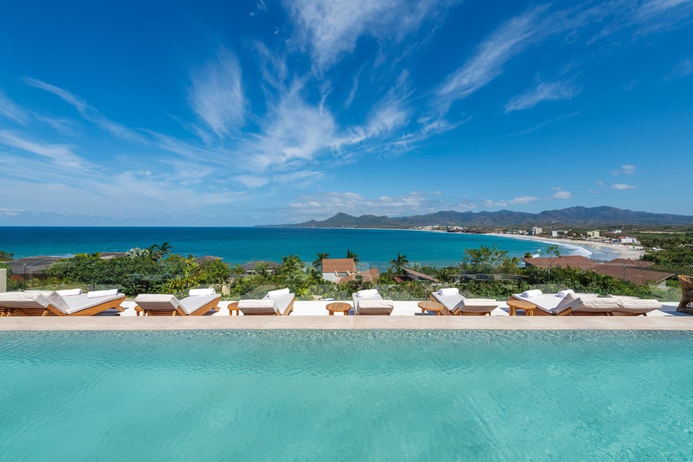 Picture yourself floating in this serene infinity pool, gazing out at pristine turquoise waters and mountain-framed coastline while lounging on plush sunbeds designed for ultimate relaxation.