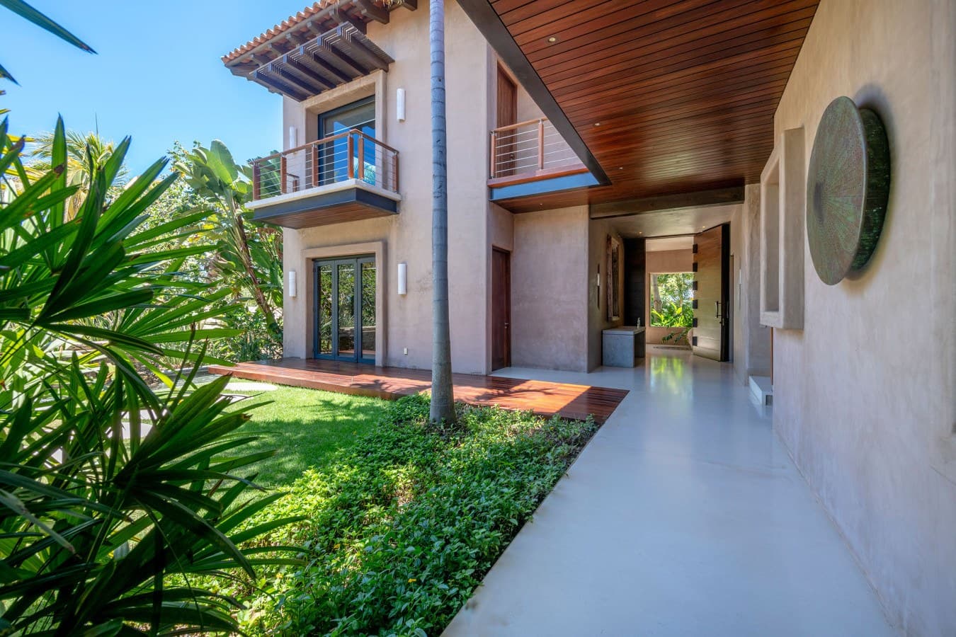 A covered walkway leads through lush tropical landscaping and manicured green lawn, connecting the main building to other areas of the property. The modern architectural design features warm wood accents and open-air elements.