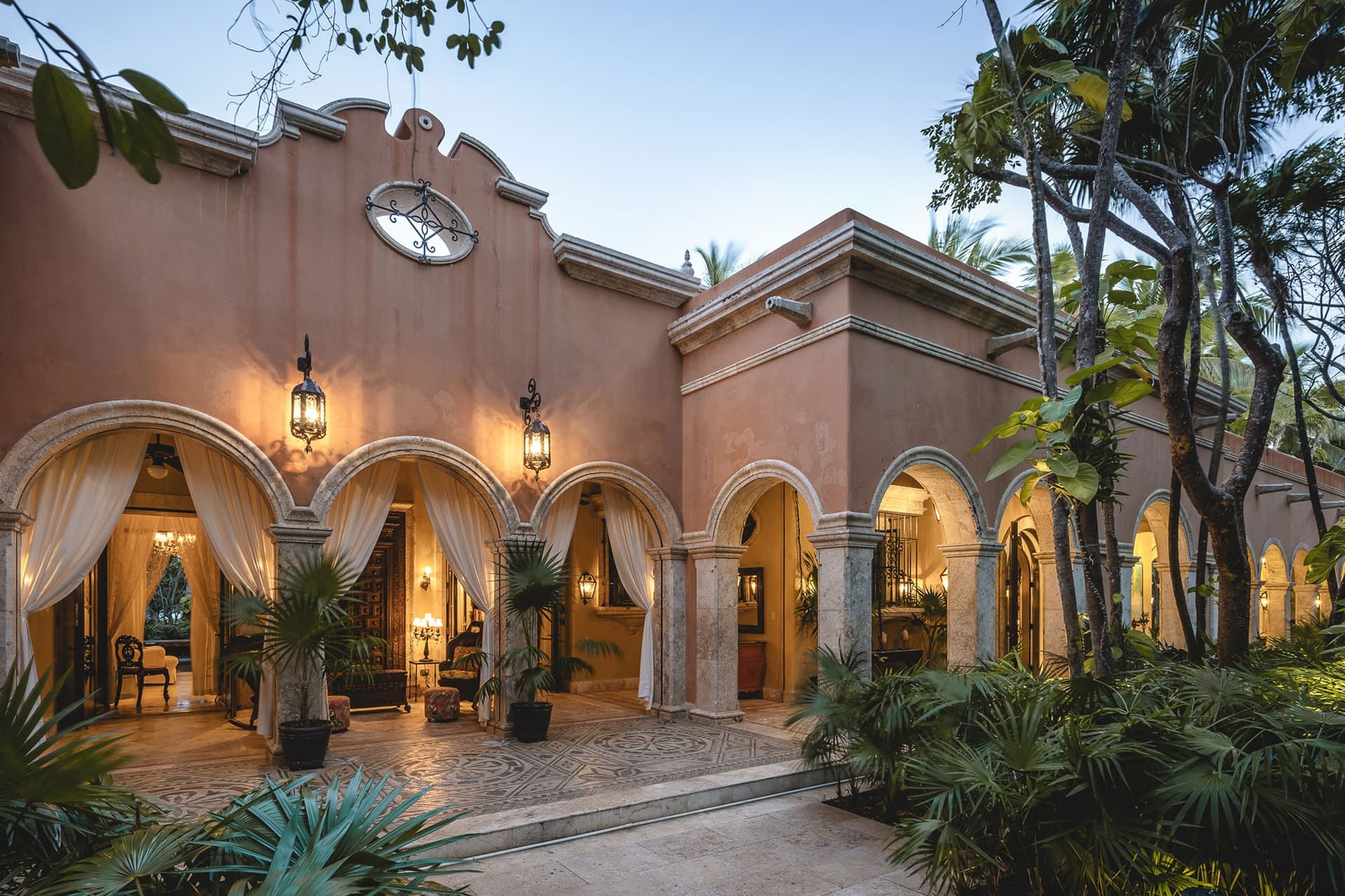 An inviting hacienda-style facade welcomes arrivals with graceful arches and warm lantern light, set amid lush tropical gardens.