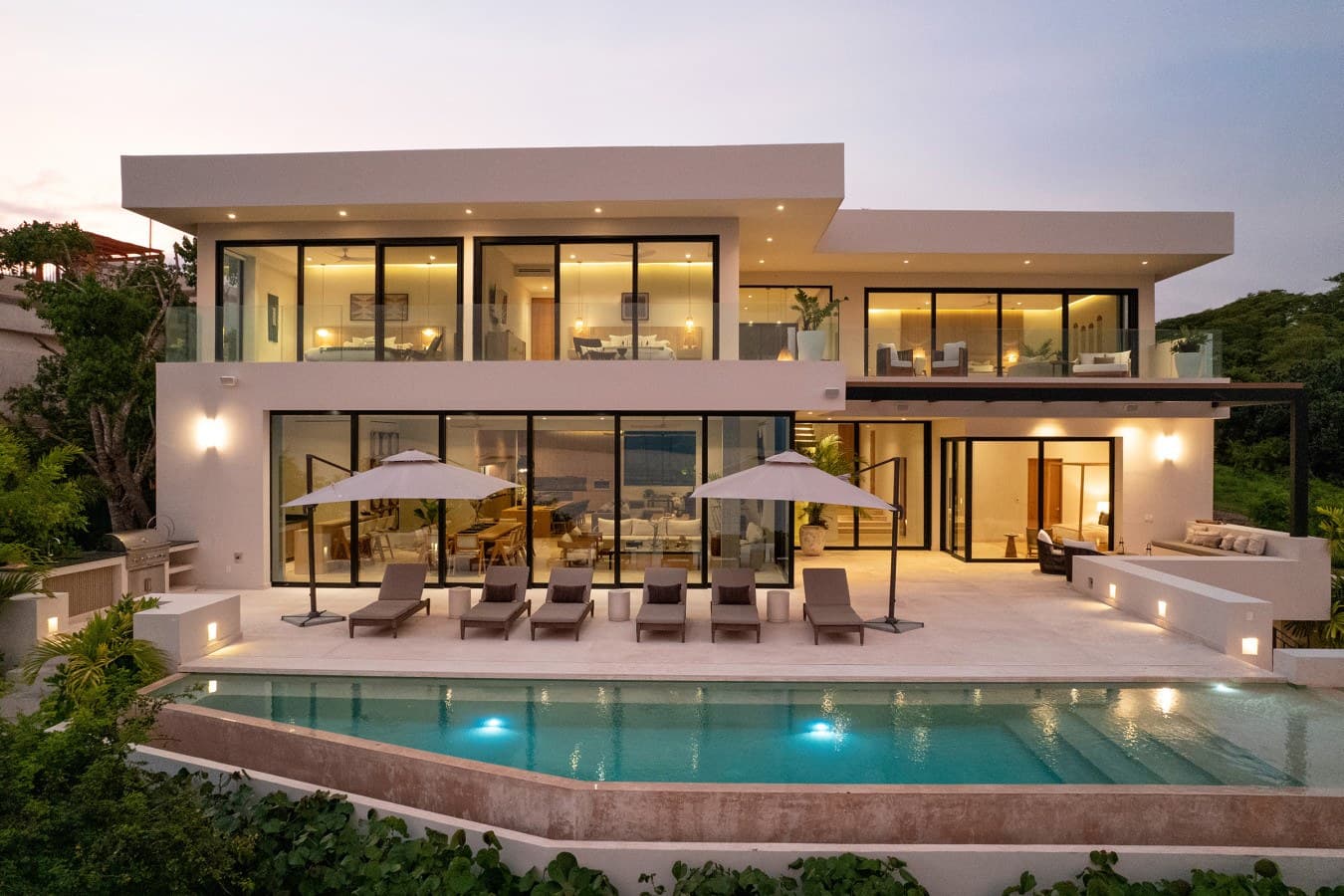 A striking contemporary villa with glowing warm interiors, expansive glass walls, and a pristine infinity pool framed by lush tropical greenery as twilight settles in.