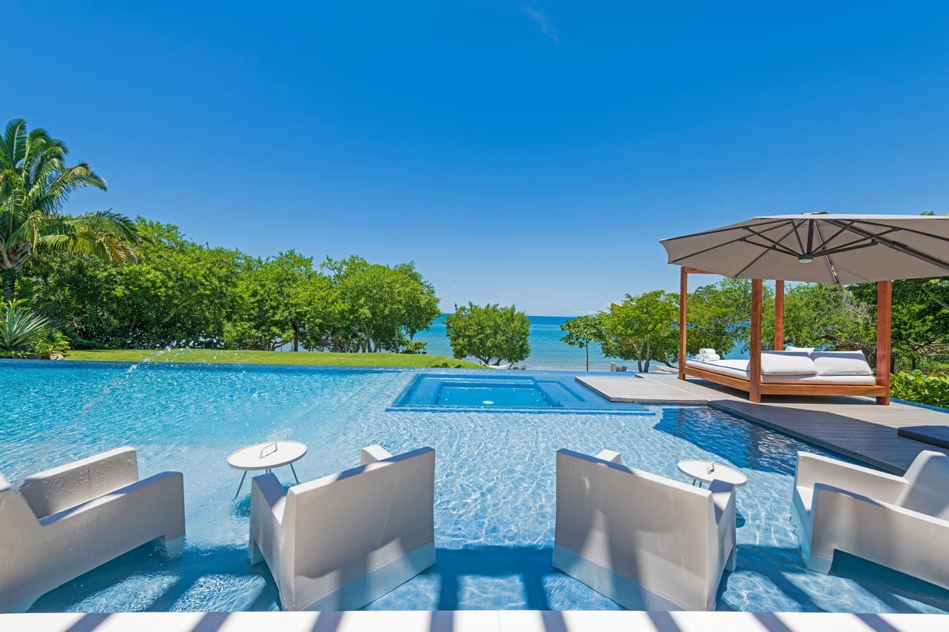 An expansive infinity pool with built-in seating and a shaded daybed overlooks lush tropical gardens and the shimmering ocean beyond under brilliant blue skies.