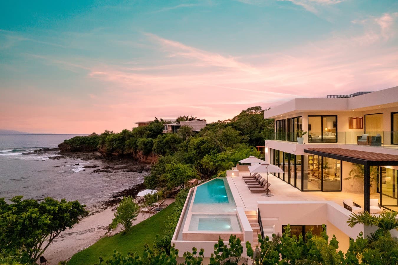 A stunning oceanfront villa perched on lush coastal cliffs, with a pristine pool deck and multiple terraces capturing the pink-hued sunset over the sea.
