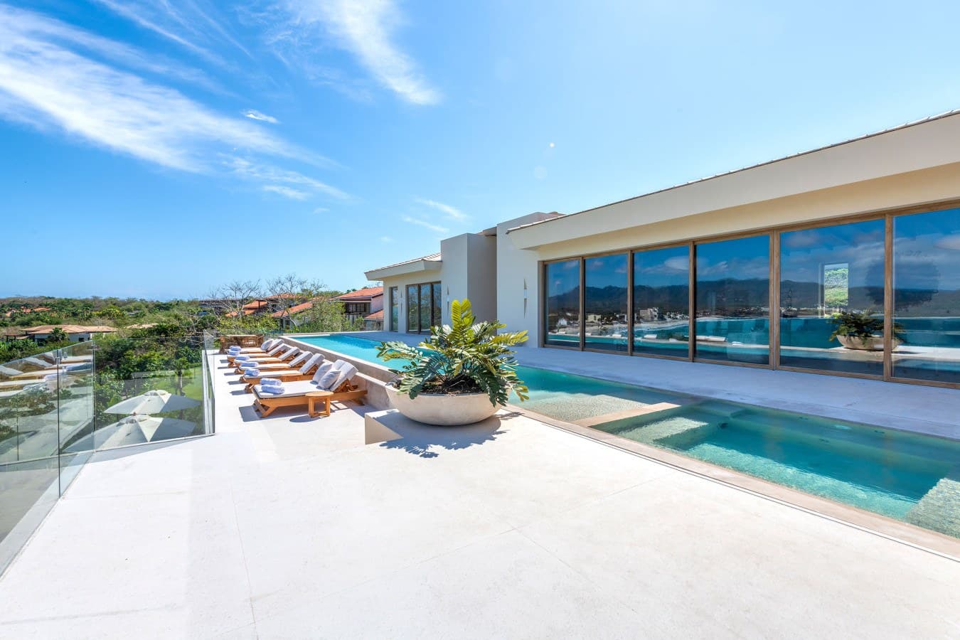 Modern luxury villa featuring an infinity pool with stunning coastal surroundings. The expansive terrace offers multiple lounge chairs for ultimate relaxation under sunny skies, surrounded by lush greenery and neighboring villas.