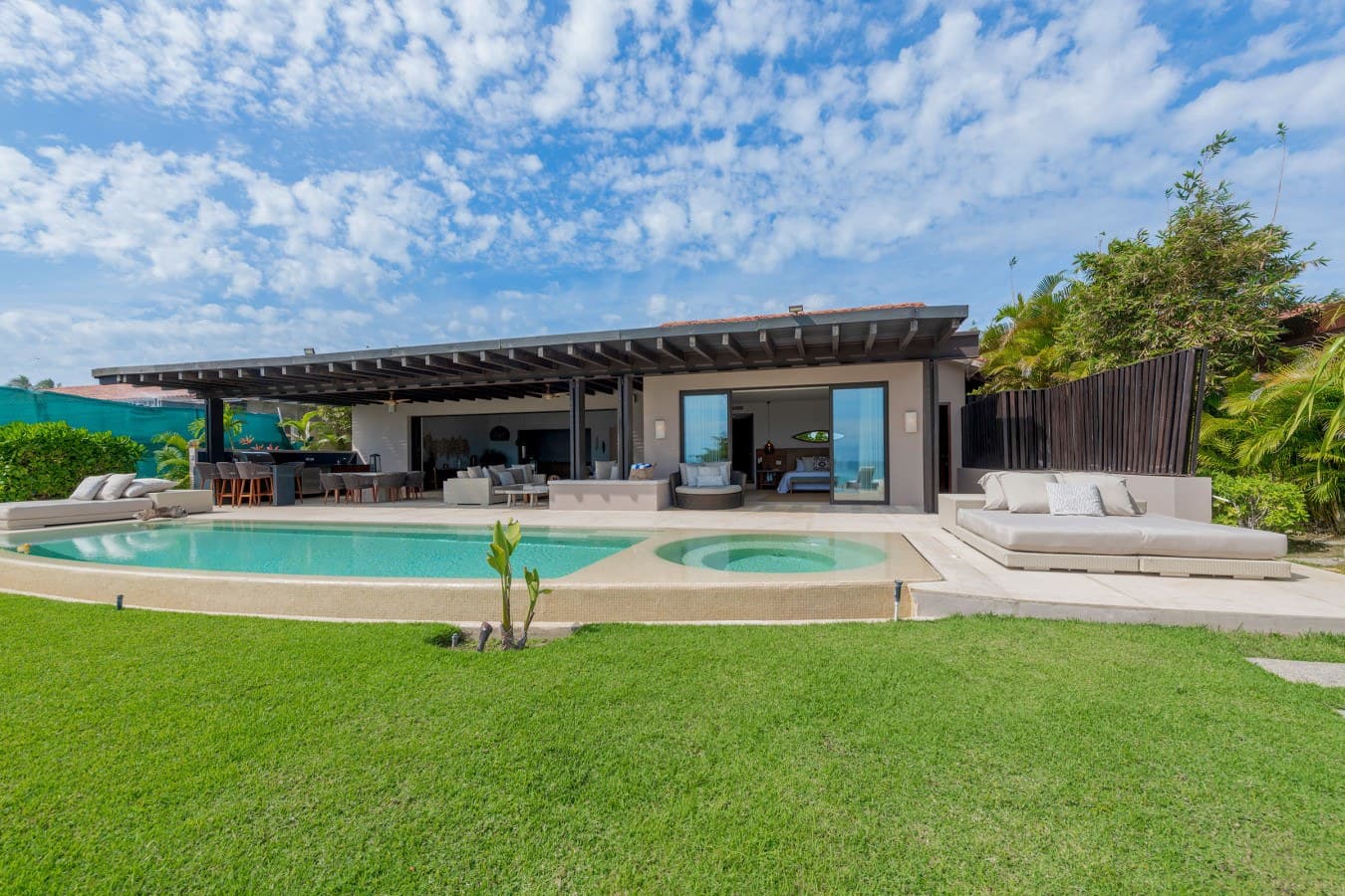 A stunning modern villa on a beautiful day, framed by lush green lawn and backed by blue skies. The property features a sparkling pool, a relaxing hot tub, and a spacious covered patio perfect for outdoor dining and lounging.