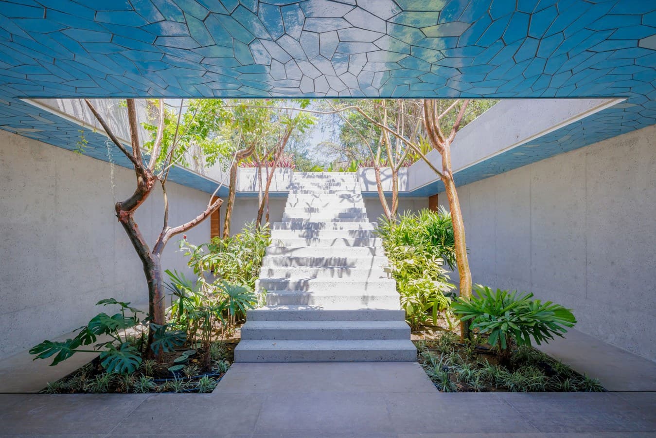 A stunning open-air entrance features a striking turquoise mosaic ceiling above white stone steps lined with lush tropical greenery and natural trees, creating an elegant welcome to the property.