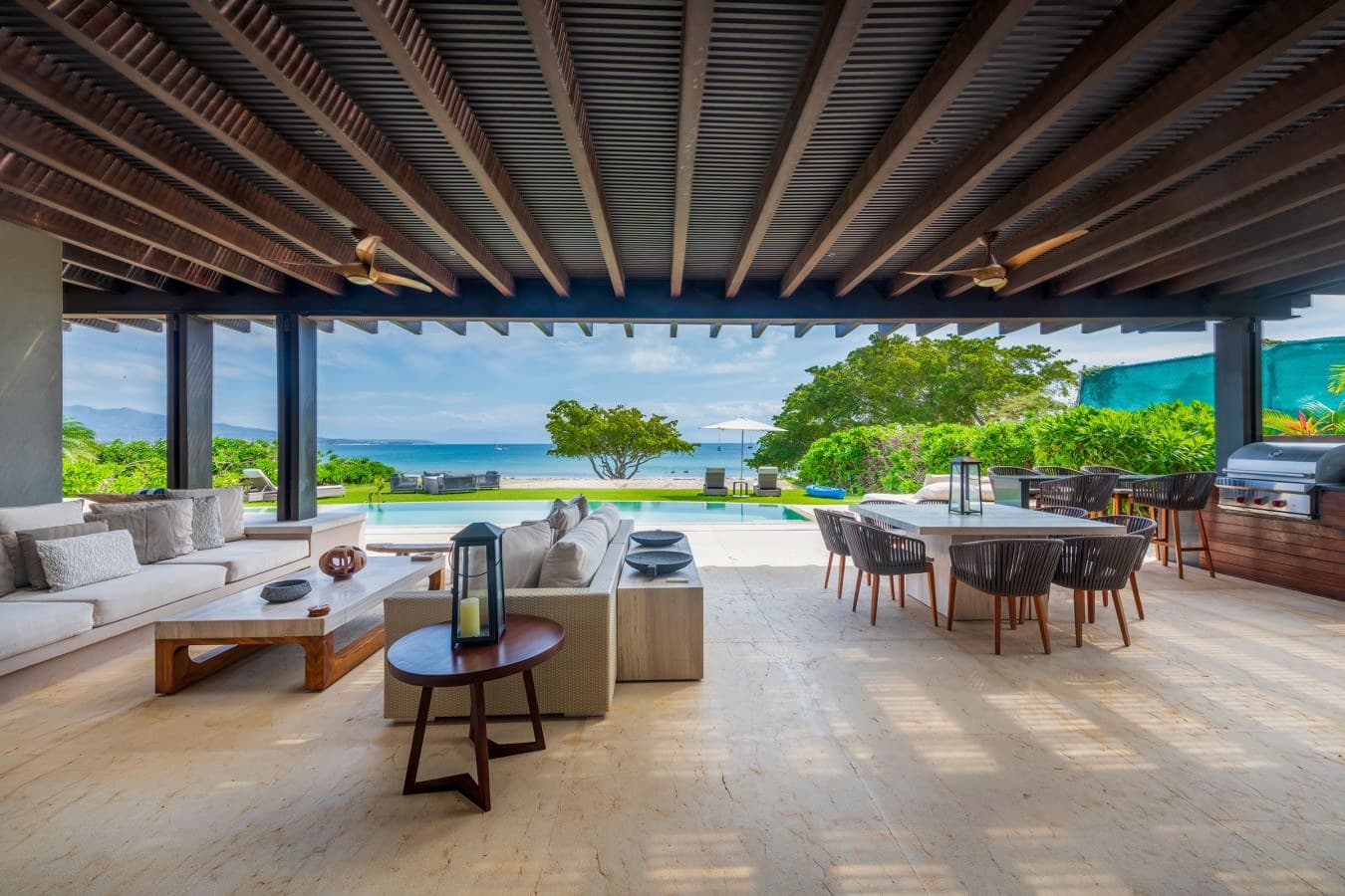 A luxurious beachfront setting featuring a covered terrace with ocean and mountain views. Modern outdoor furnishings blend seamlessly with lush tropical surroundings and a pristine pool area.