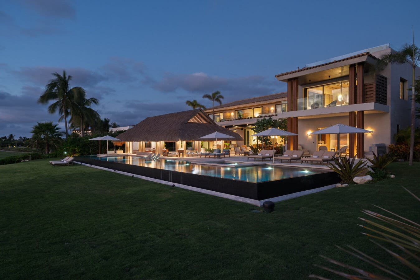 A stunning modern villa glows warmly at twilight, showcasing its expansive infinity pool, elegant outdoor lounging areas, and lush tropical gardens beneath a painted evening sky—your private paradise awaits.