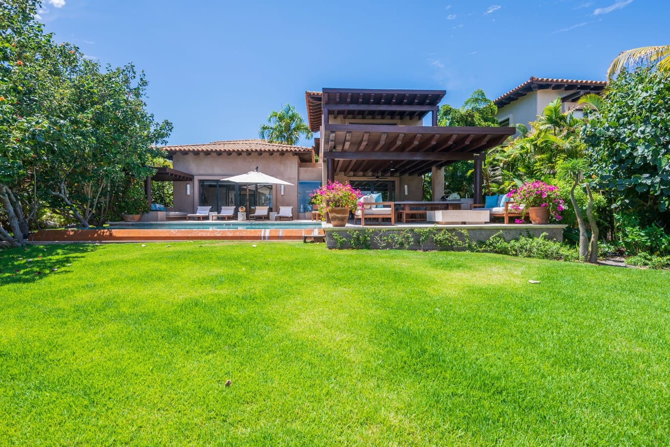 This beautiful villa features a sprawling lawn surrounded by lush gardens and tropical palms, with a spacious covered terrace and private pool perfect for outdoor living.