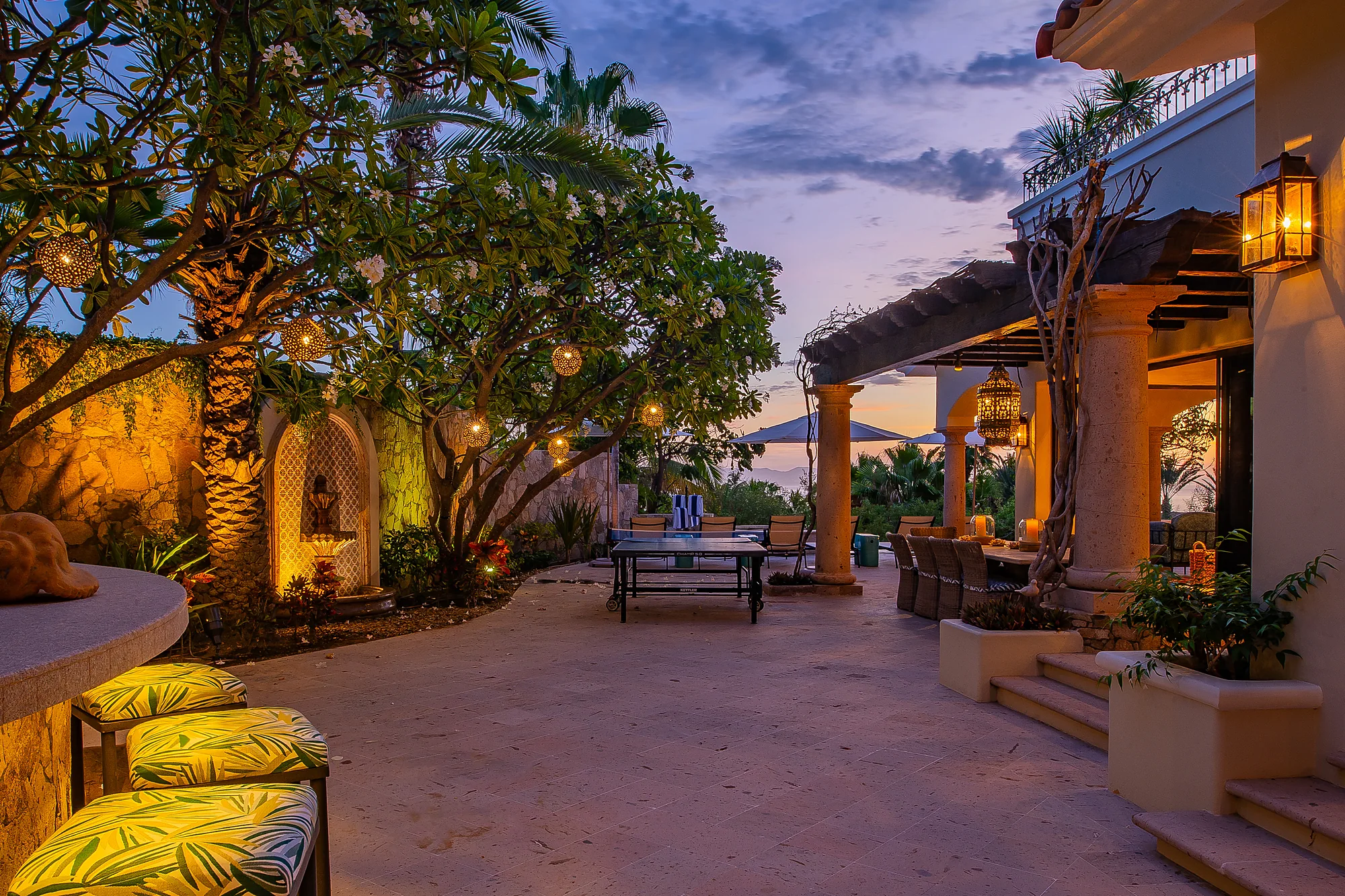 This outdoor courtyard glows as dusk settles, with lanterns casting warm light across inviting seating areas, lush palms, and serene architecture—an enchanting space for evening relaxation.
