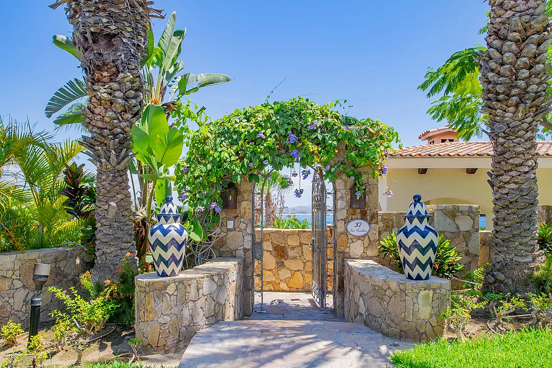 The property entrance features a vine-draped stone archway flanked by tropical palms and decorative blue-and-white vases, welcoming guests to a Mediterranean-inspired coastal retreat.