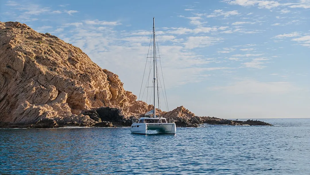 A white catamaran anchored in crystal-clear waters beside dramatic golden cliffs under a partly cloudy sky.