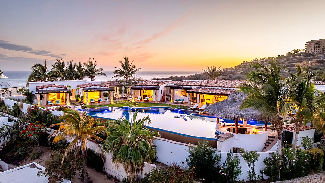 Luxurious beachfront villa with infinity pool overlooking the ocean, surrounded by lush tropical gardens and mountains at sunset.