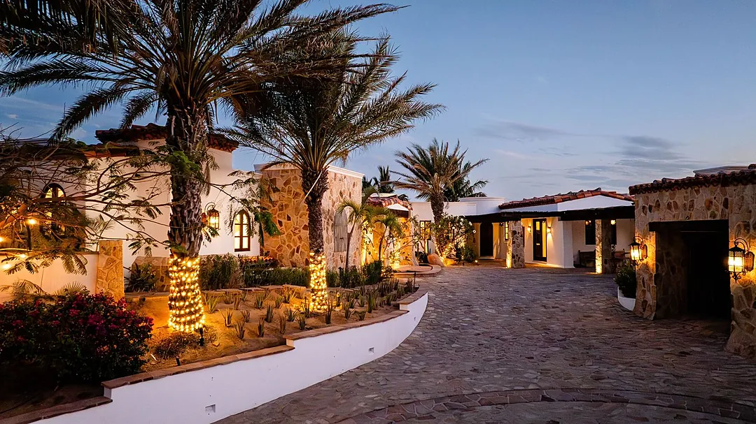 A beautifully landscaped property exterior showcasing Spanish-style architecture with stone accents, palm trees, and ambient lighting creating an inviting entrance at dusk.