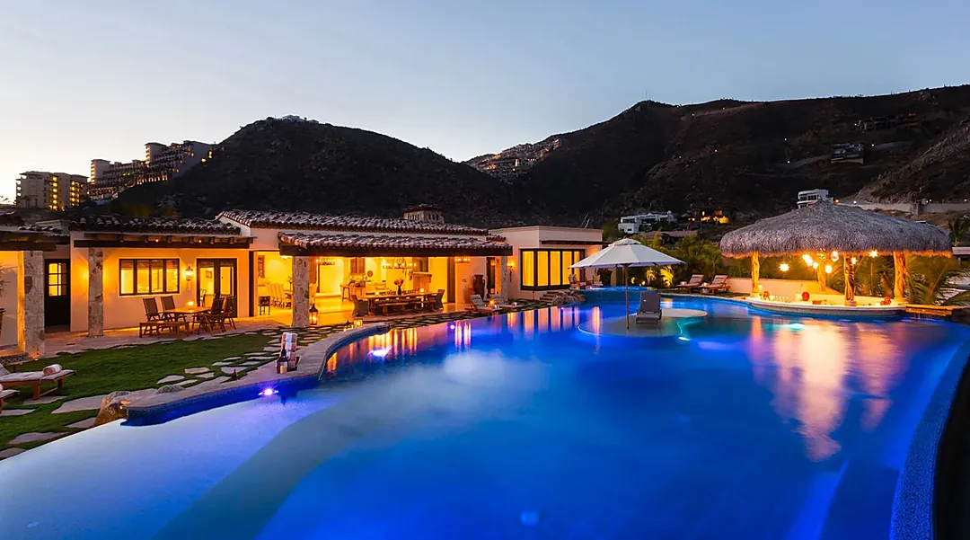 A spectacular hillside property with a stunning illuminated pool nestled between mountains. The spacious villa features covered outdoor dining areas, tropical palapas, and ambient evening lighting throughout.