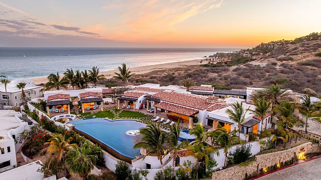 An aerial view of a beachfront property at sunset, with a swimming pool, palm trees, and hillside terrain meeting the ocean. The property features white stucco architecture and offers coastal landscape views during golden hour.