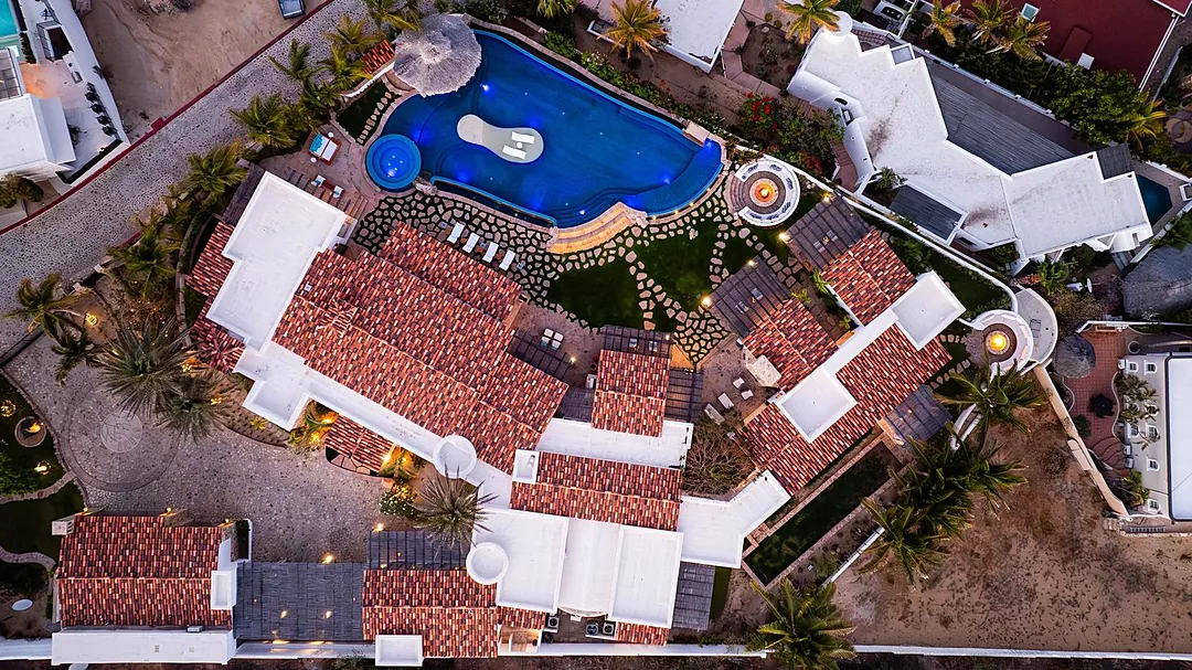 Aerial view showcasing the property's distinctive architecture, terracotta roofing, blue-lit swimming pool, and lush palm-studded grounds nestled within the surrounding neighborhood.