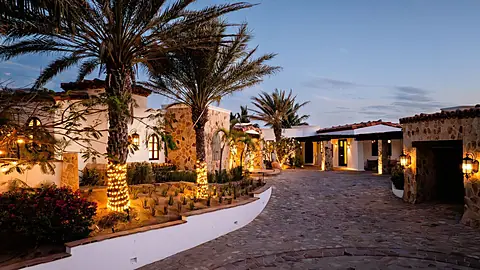 A beautifully landscaped property exterior showcasing Spanish-style architecture with stone accents, palm trees, and ambient lighting creating an inviting entrance at dusk.