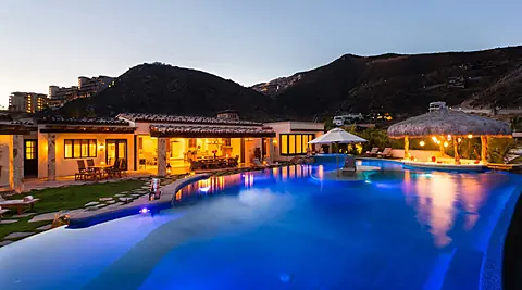 A spectacular hillside property with a stunning illuminated pool nestled between mountains. The spacious villa features covered outdoor dining areas, tropical palapas, and ambient evening lighting throughout.