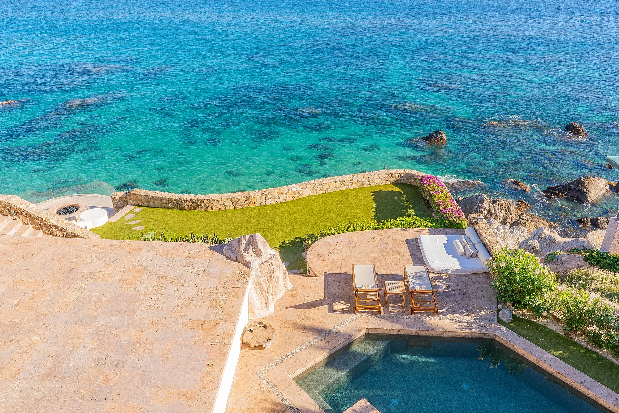 An aerial view showcasing the property's coastal setting with a private pool overlooking pristine turquoise waters and rocky shoreline, surrounded by lush landscaped grounds.
