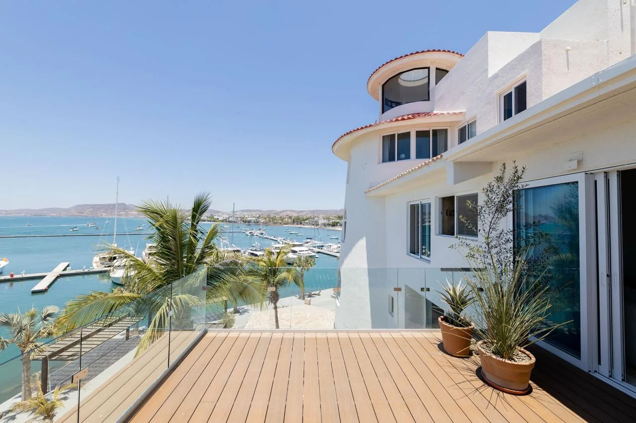 Stunning waterfront property features a Mediterranean-style white exterior with terracotta roof and curved architecture overlooking a serene marina with boats and palm trees.