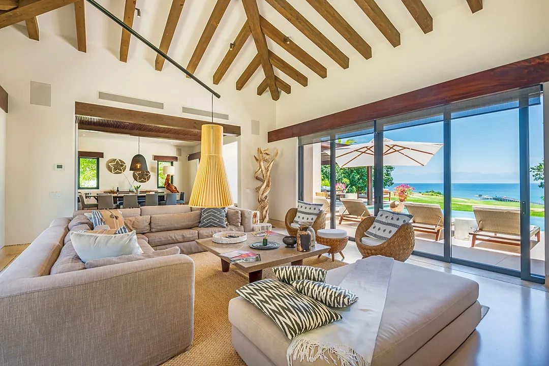 Step into your sunlit sanctuary where dramatic wooden beams soar overhead and floor-to-ceiling windows frame endless ocean views. Sink into plush seating as sea breezes drift through open doors to your private terrace.