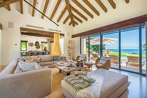 Step into your sunlit sanctuary where dramatic wooden beams soar overhead and floor-to-ceiling windows frame endless ocean views. Sink into plush seating as sea breezes drift through open doors to your private terrace.