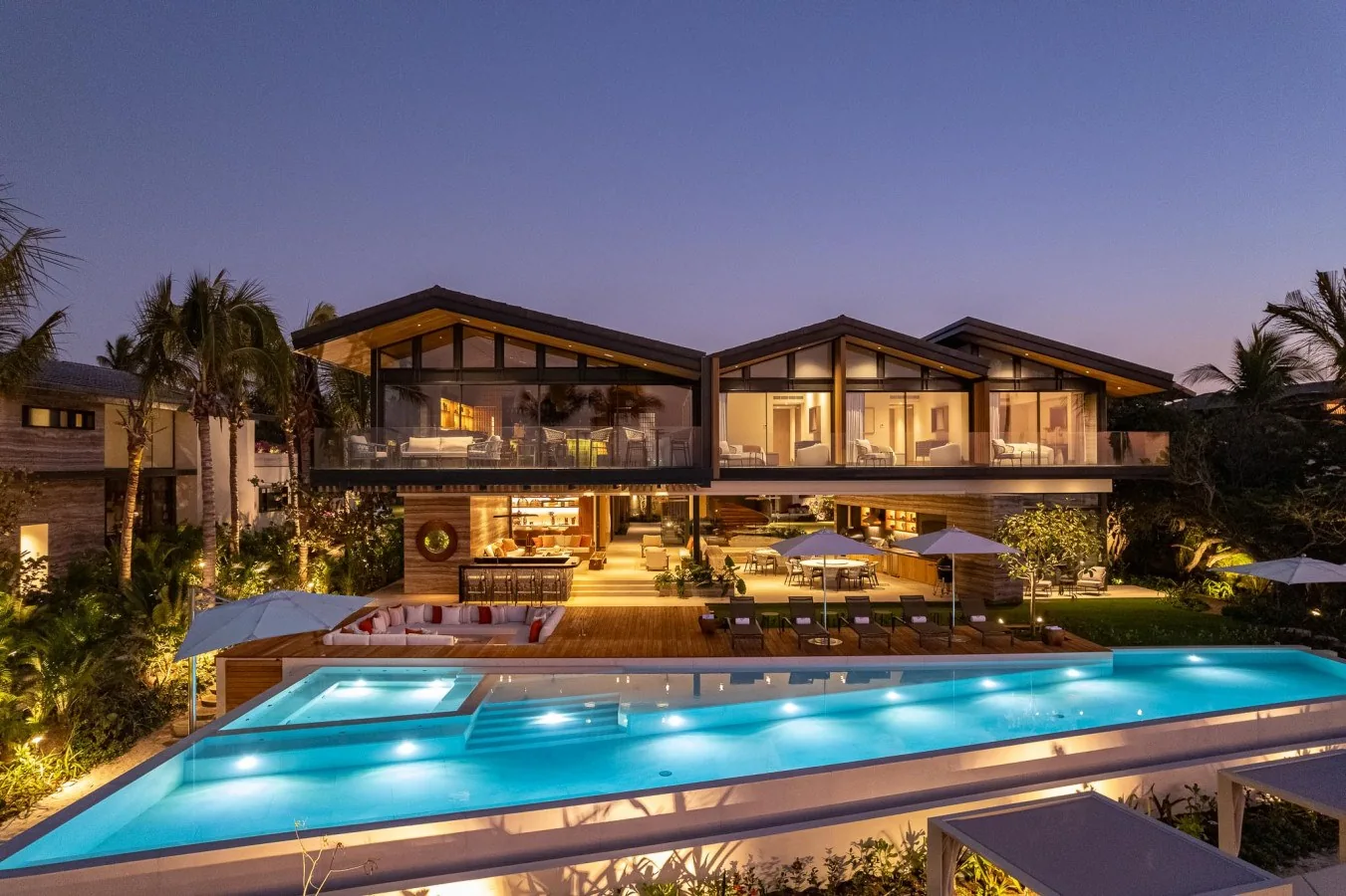 A stunning beachfront villa illuminated at twilight, surrounded by swaying palms and tropical landscaping, with a dramatic blue swimming pool at its heart.