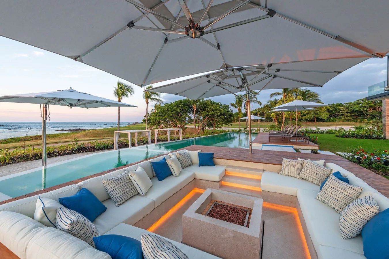 A stunning beachfront outdoor lounge invites you to unwind. Sink into plush seating around a glowing fire feature as the sun sets over the ocean—pure coastal paradise awaits.