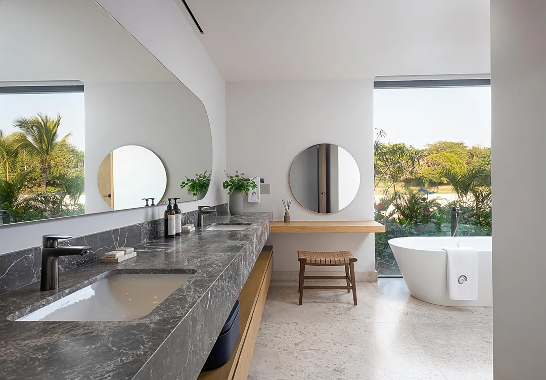 Bathroom 1 (Master ensuite) · Step into your private sanctuary where sleek marble counters meet natural wood, and a freestanding soaking tub awaits your relaxation. Large windows frame lush tropical greenery, bringing the outdoors in.