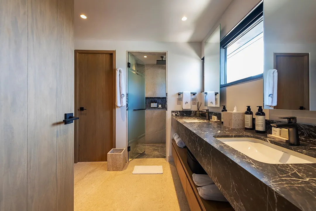 Bathroom 2 · Step into your private spa-like retreat with luxurious dual sinks set in elegant dark marble, soft robes waiting for you, and a sleek glass-enclosed shower just beyond.