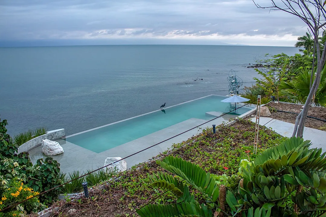 A dramatic infinity pool set into lush tropical gardens, where the horizon blurs seamlessly into the ocean beyond and clouds paint moody patterns across an endless sky.